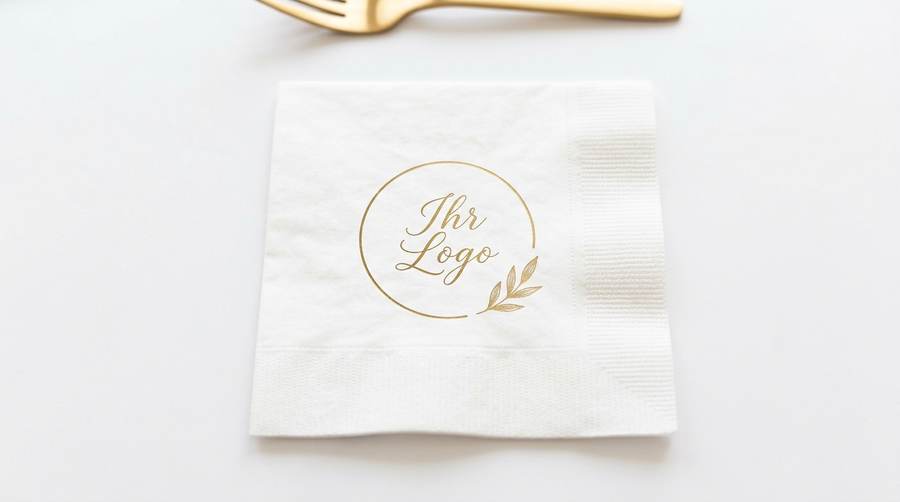 Weiße Papierserviette mit goldenem Rundlogo 'Ihr Logo' und Blattmotiv, daneben goldene Gabel